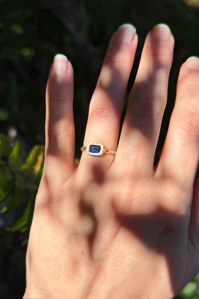 Hand wearing a gold ring with a blue gemstone against a blurred natural background