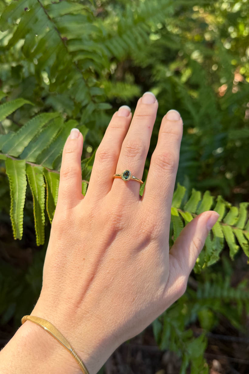 Hand wearing a gold ring with a green gemstone against a fern background