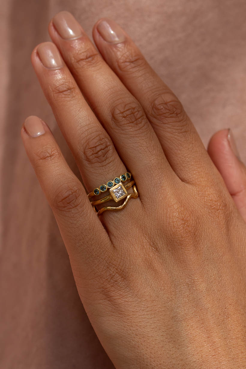 Hand wearing a stack of three yellow gold rings: the Gumnut Wedding Band with teal Australian sapphires, the Clover Ring with a white diamond, and the Tendril Ring in recycled gold