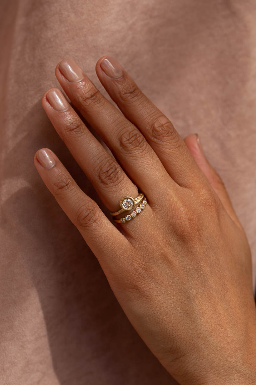 Hand wearing a stack of gold rings: the Honeysuckle Ring, a textured solitaire engagement ring with a champagne diamond, and the Gumnut Wedding Band with diamonds