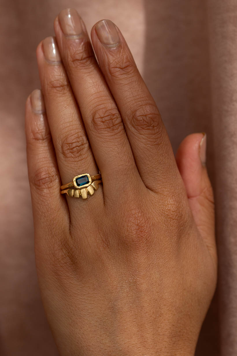 A gold ring with a rectangular blue stone and a sunburst design on the band, shown worn on a person's ring finger against a soft brown background. The Arbor ring with an Australian deep blue emerald cut sapphire set in textured bezel style stacked with he texture Keystone ring