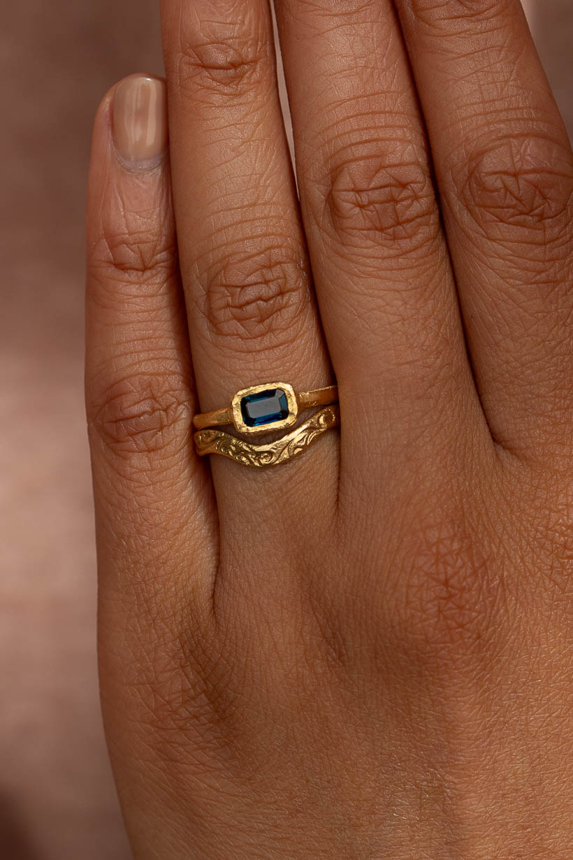 Arbor engagement ring with a deep blue emerald cut Australian sapphire in a textured bezel, stacked with the Whisper curved floral engraved wedding band on the hand