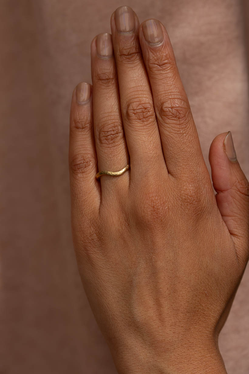 A close-up of a hand wearing a simple curved gold band ring against a warm brown background. The 2mm wide Breeze ring, a textured, organic, handcrafted wedding band made in recycled gold in Melbourne.