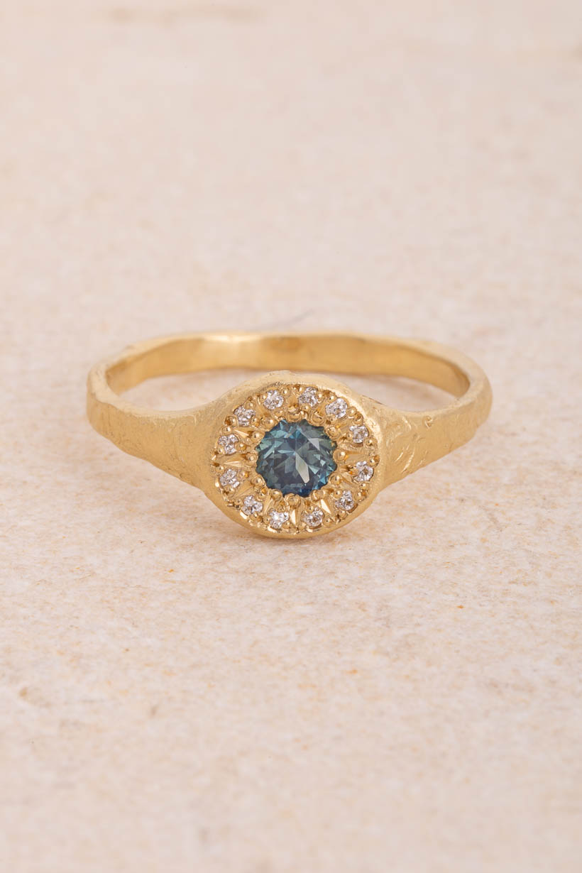 Floral Relic Halo Signet engagement ring with an ethically sourced light blue Australian sapphire in a halo of 16 star-set white diamonds, on a floral-engraved signet band handmade in Melbourne from recycled gold, front view on limestone