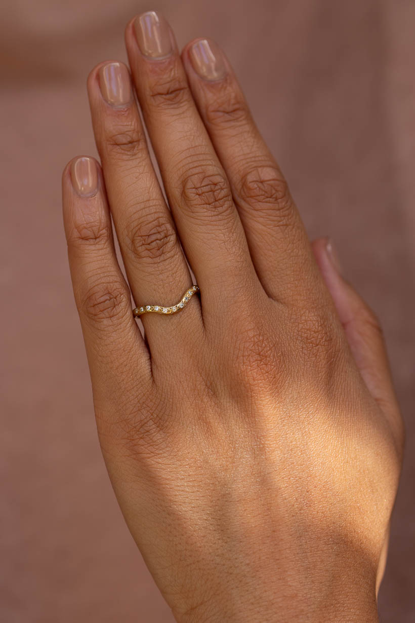 A close-up of a hand wearing a delicate gold ring, the rosette ring, a textural, organic, gold curved wedding ring with a row of small white diamonds, set against a soft, warm brown background.