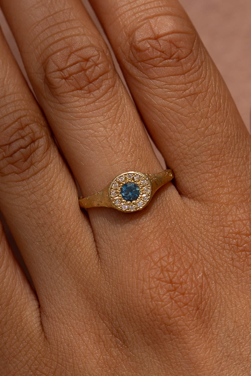 A textured gold signet ring with a round blue Australian sapphire surrounded by a halo of small natural white diamonds shown worn on a hand.