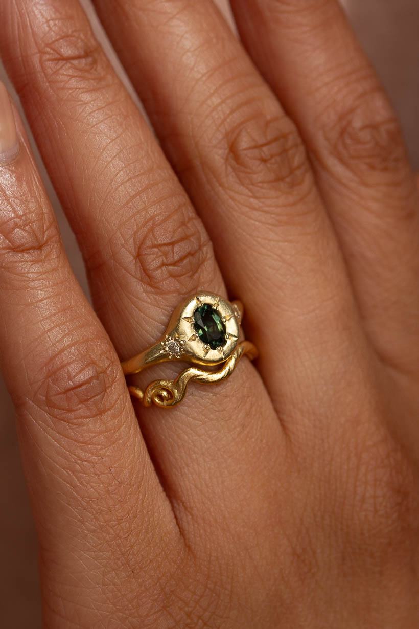 Close-up on hand of Monet's Garden signet style engagement ring with oval green Australian sapphire details showing flower-set champagne diamonds in hand-textured gold. Stacked with curved vine wedding ring in recycled yellow gold.