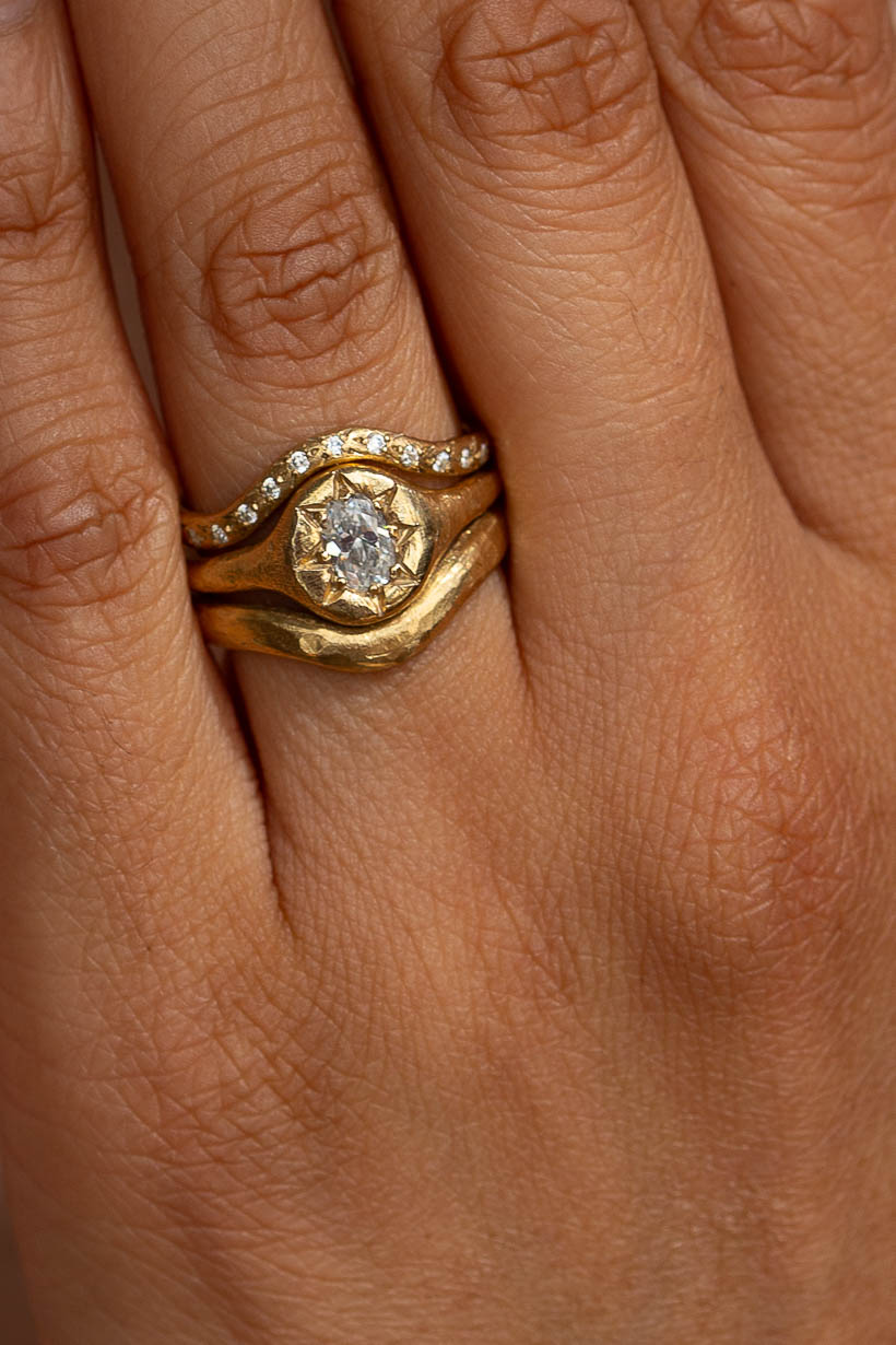 Close up of Monet's Garden oval diamond engagement ring in recycled yellow gold worn on hand stacked with curved Rosette ring with white diamonds wedding band and 3mm Breeze curved wedding band