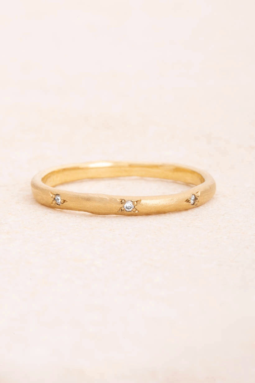 Rosette ring with three natural white diamonds in petal-like rosette settings on a fine textured recycled gold band, flat lay, Ada Hodgson