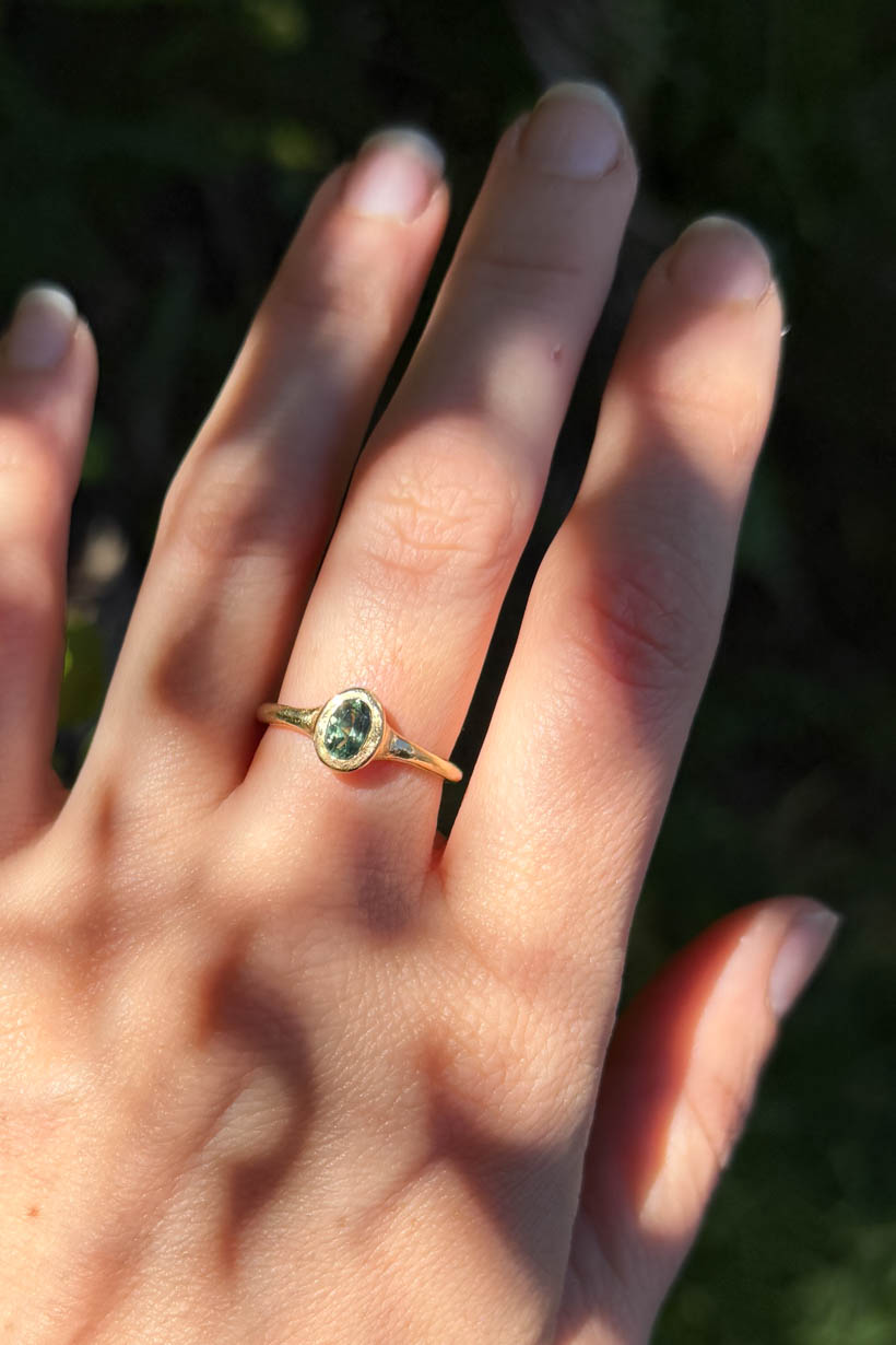 Hand wearing a gold ring with a green gemstone against a blurred natural background