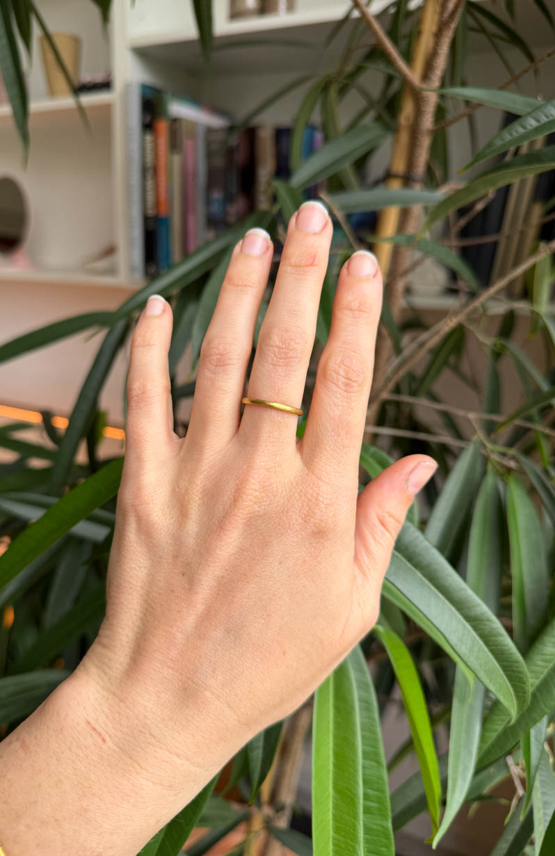 Hand wearing a gold ring with a blurred indoor setting. 2mm wide softly textured and organic breeze ring. 