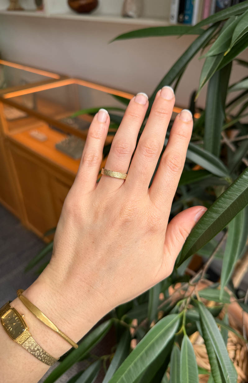 Hand wearing a textured gold wedding ring with a plant in the background
