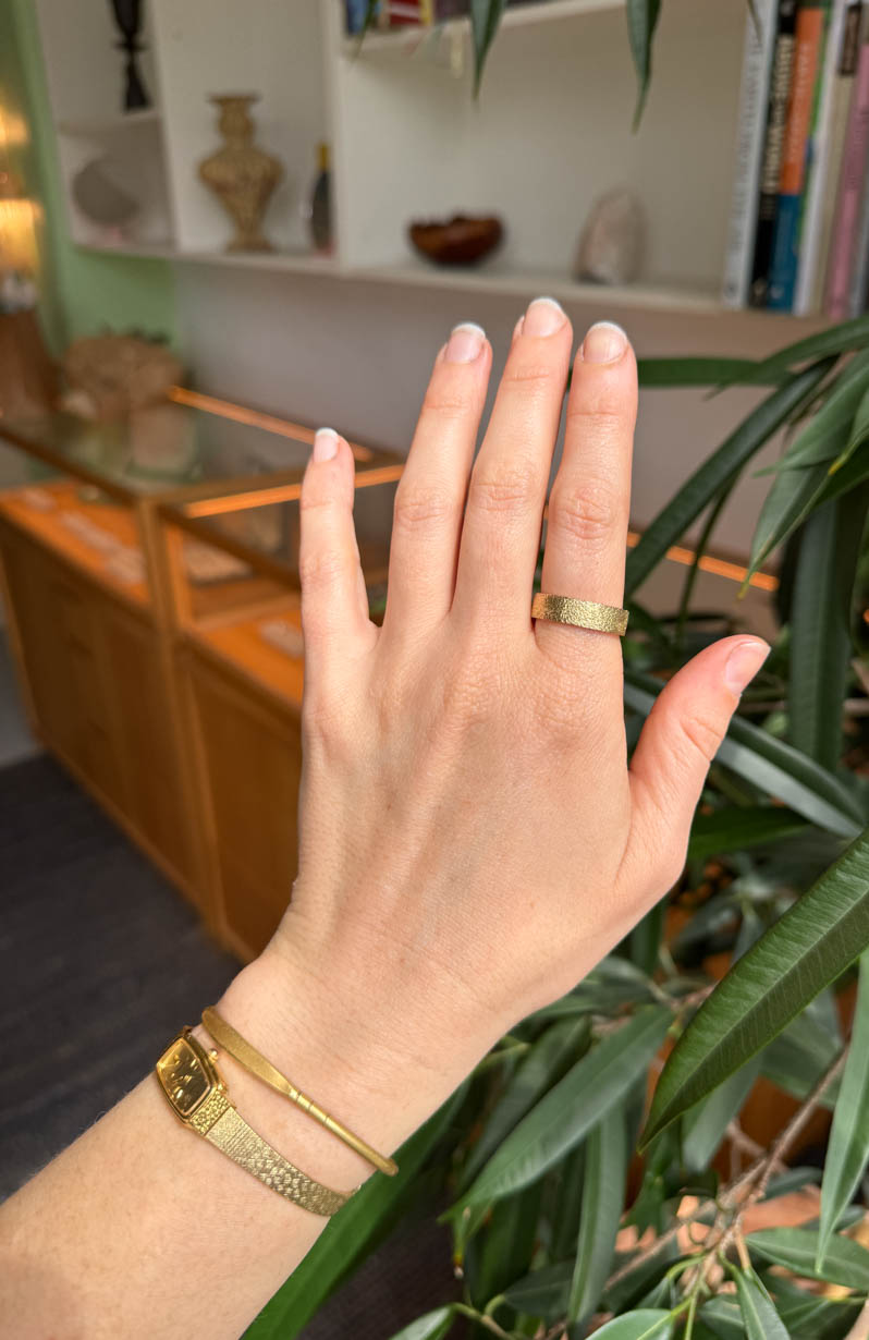 Hand wearing a gold ring and bracelet with a blurred indoor background. Textured gold wedding band. Citrus ring. 