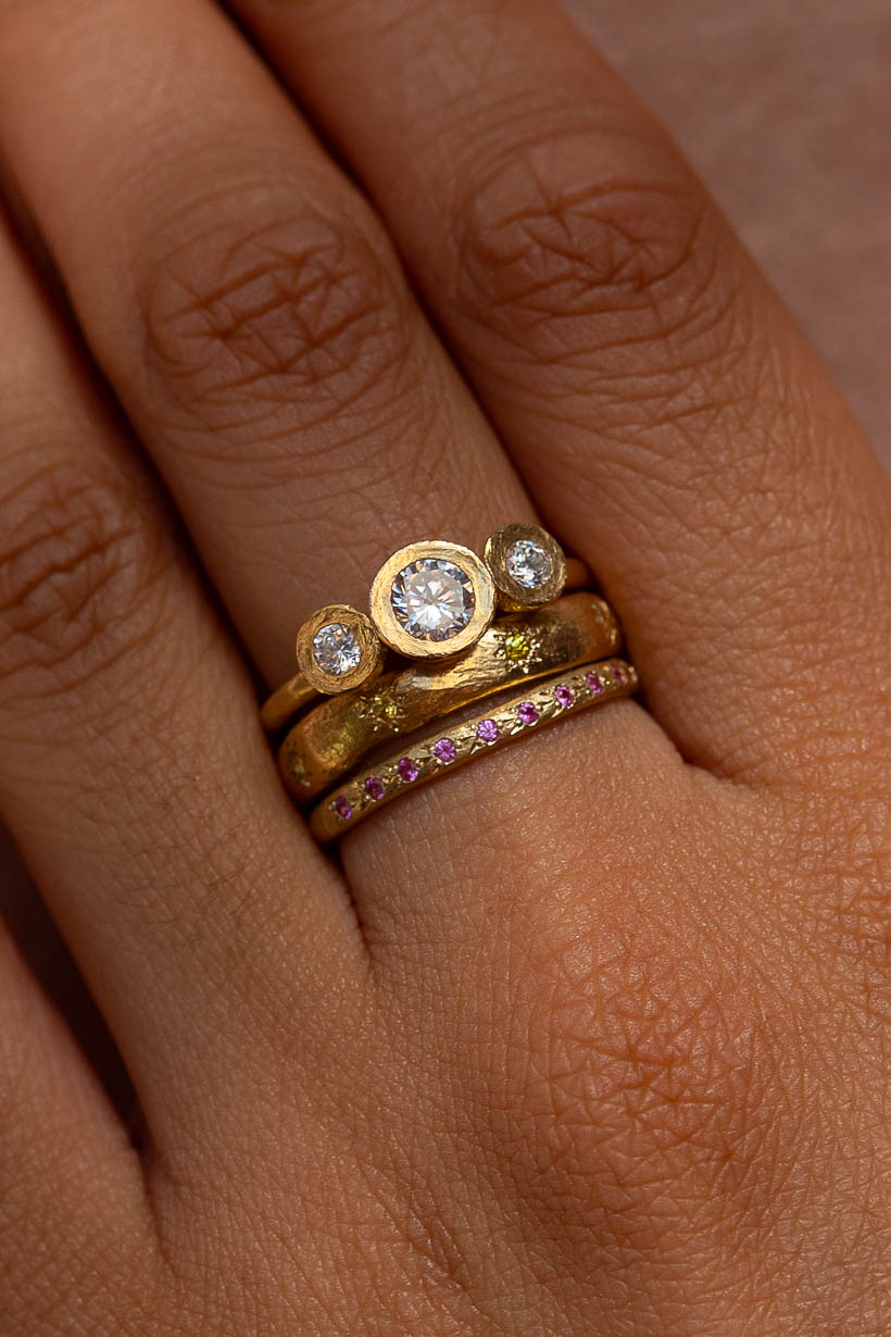 Close-up of a stacked yellow gold ring trio: the Gumnut Trilogy Ring with diamonds, the Daisy Chain Ring with yellow sapphires, and the Rosette Ring with pink sapphires