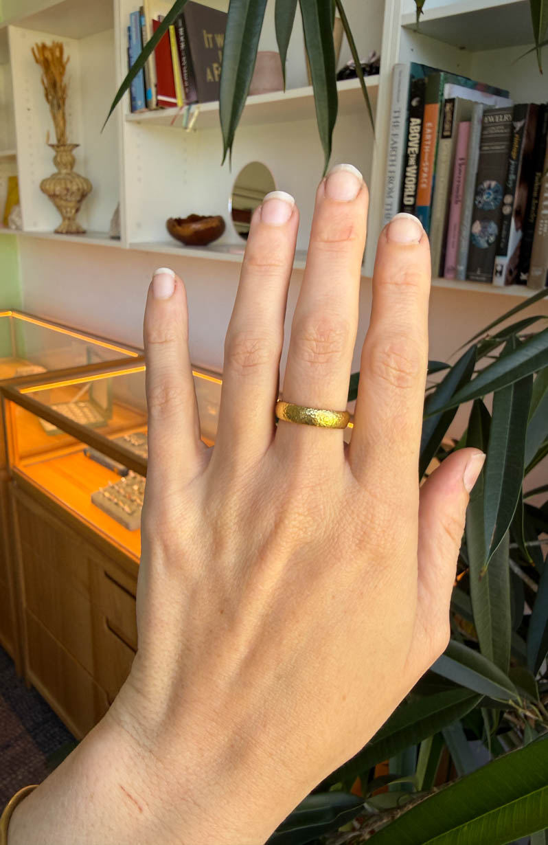 Hand wearing a gold ring with a blurred indoor setting in the background