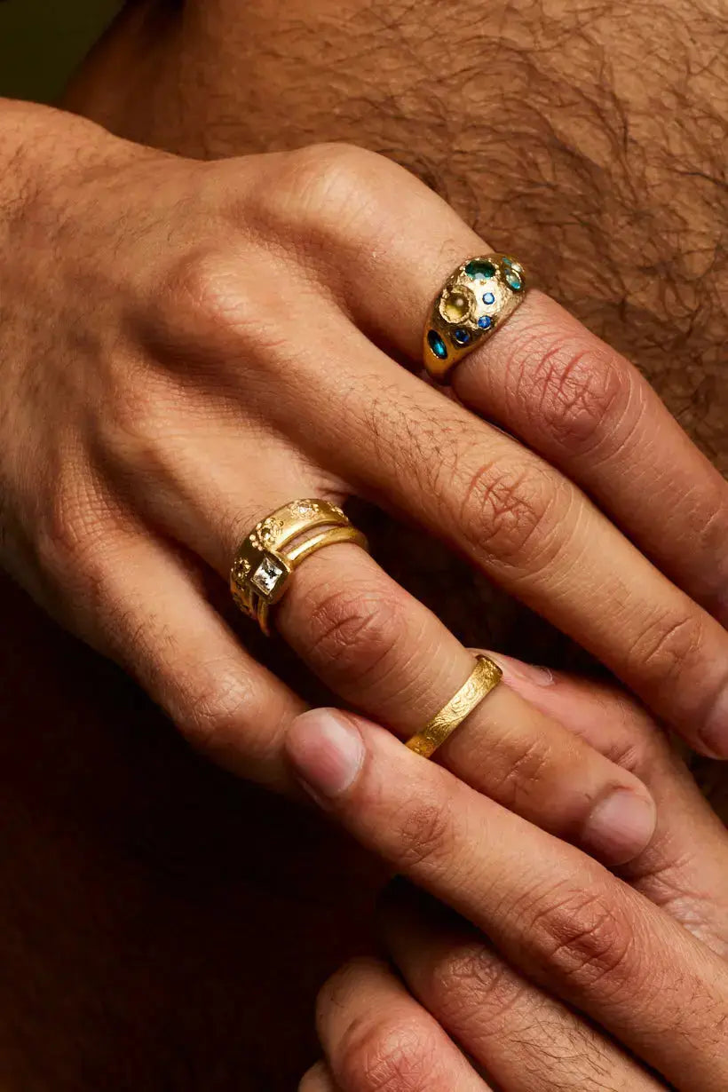 The Rockpool ring, a wide gold wedding band set with scattered blue and green precious gemstones styled on models hand with other band bands and diamond engagement rings. 