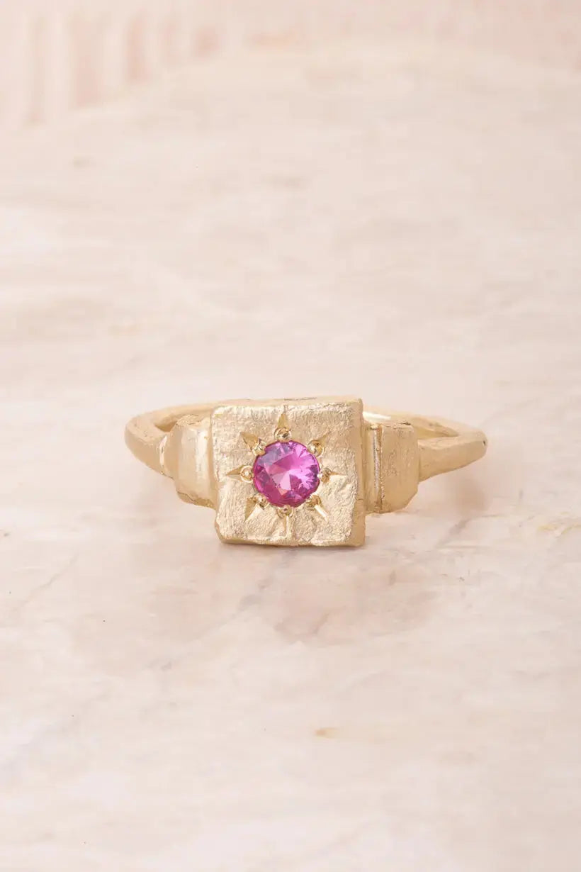 Flatlay of the Dawn Signet ring in recycled gold, set with a vibrant pink sapphire, softly textured and hand-carved.