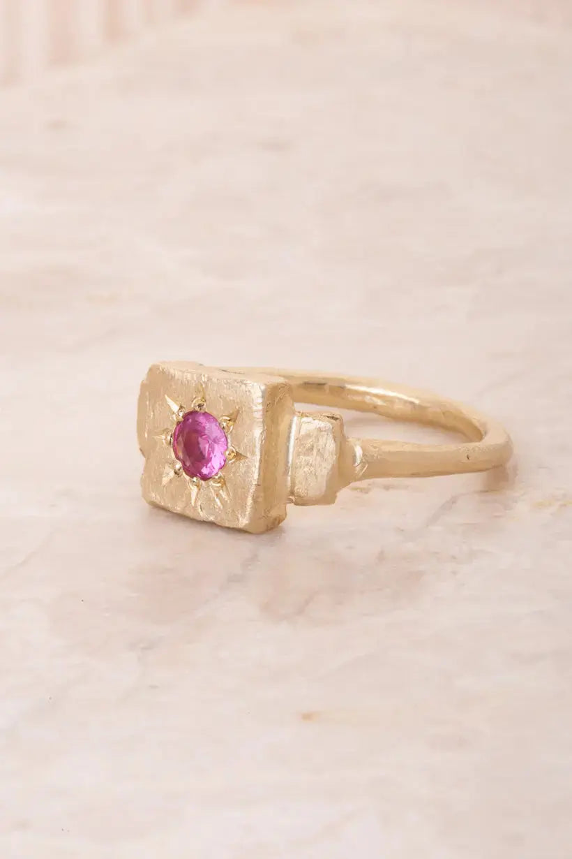 Side view flatlay of the Dawn Signet ring, showing its irregular hand-carved band and bright pink sapphire centre.