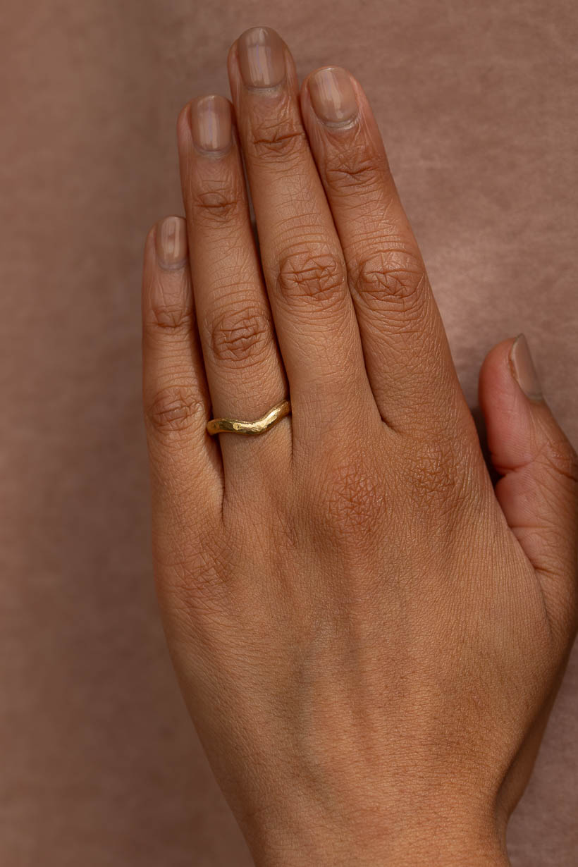 A close-up of a hand wearing a simple, textured curved gold band ring against a solid warm brown background. The Breeze curved wedding band in recycled, textured, organic gold, 3mm wide. Handmade in Melbourne by jeweller Ada Hodgson