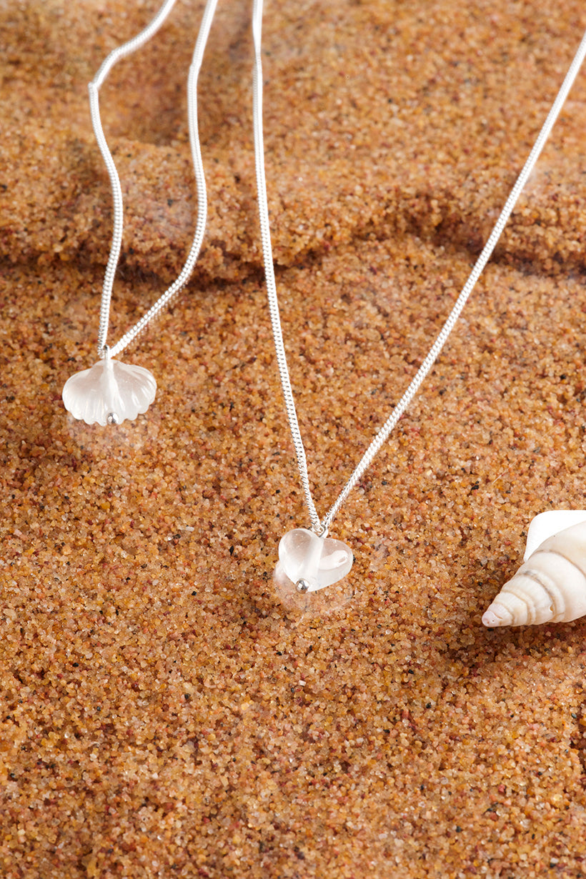 Glass shell and heart pendants on necklaces lying on sand with shells nearby