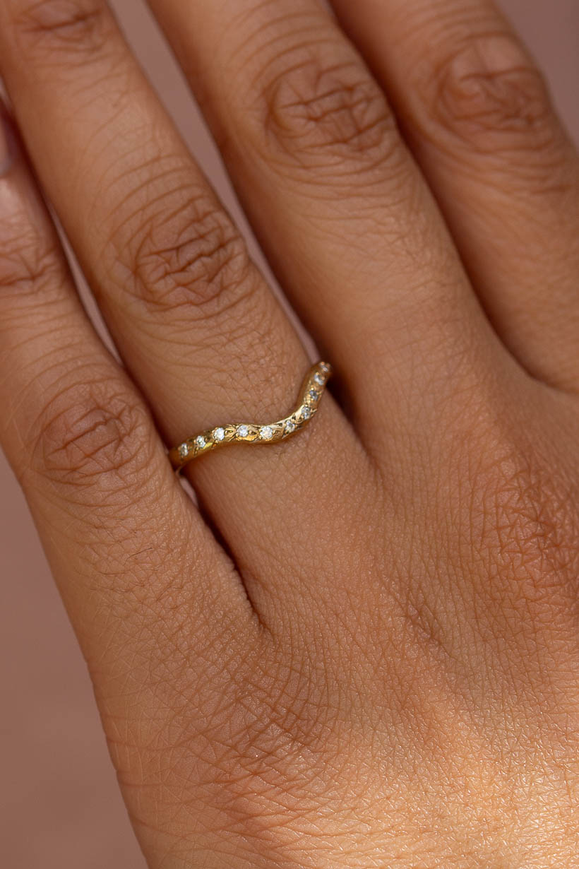 A close-up of a hand wearing a delicate wavy yellow gold ring, the curved rosette ring in organic, textural recycled gold with natural diamonds