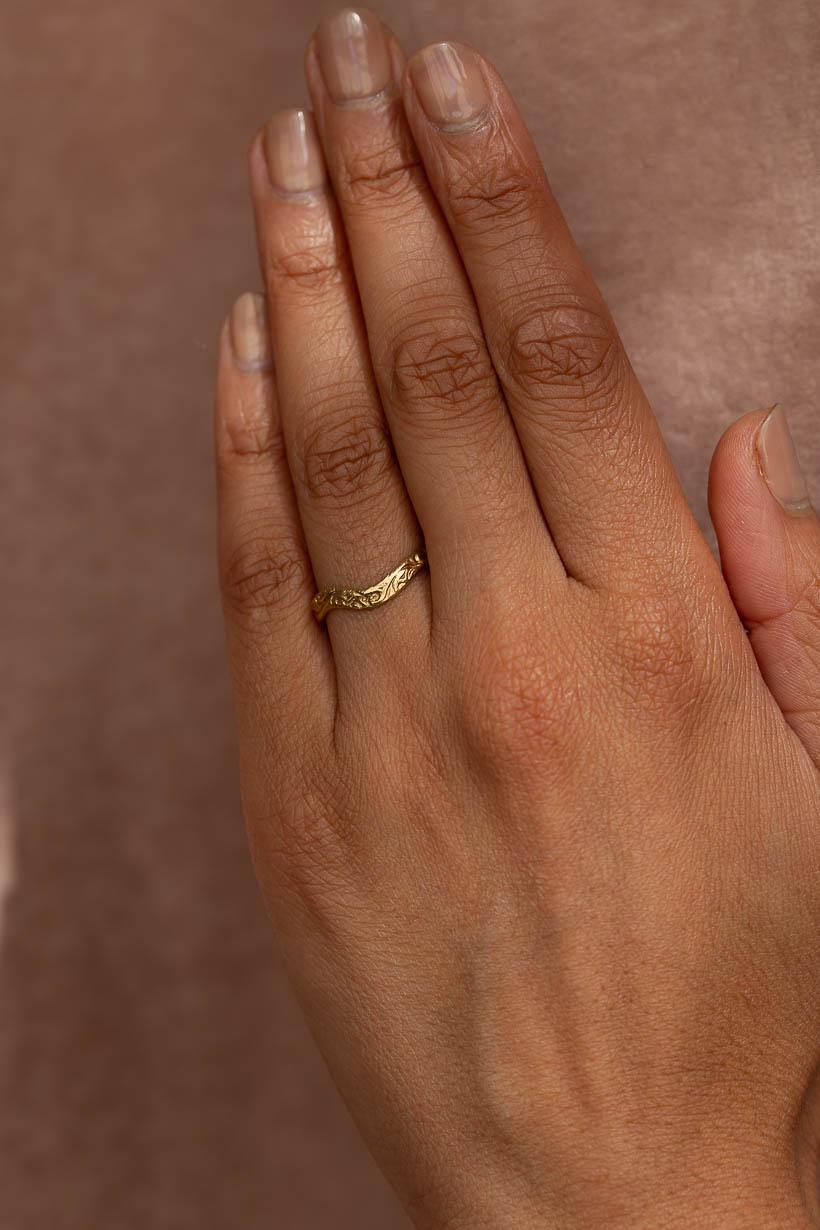 A close-up of a person's hand wearing a wide engraved  textured gold band ring on the ring finger, the whisper ring in recycled gold. 