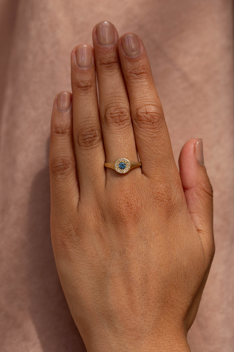 A close-up of a hand wearing a gold ring, the Floral Relic Halo Signet engagement ring with a central round blue Australian sapphire surrounded by small natural diamonds, set against a soft pink fabric background.