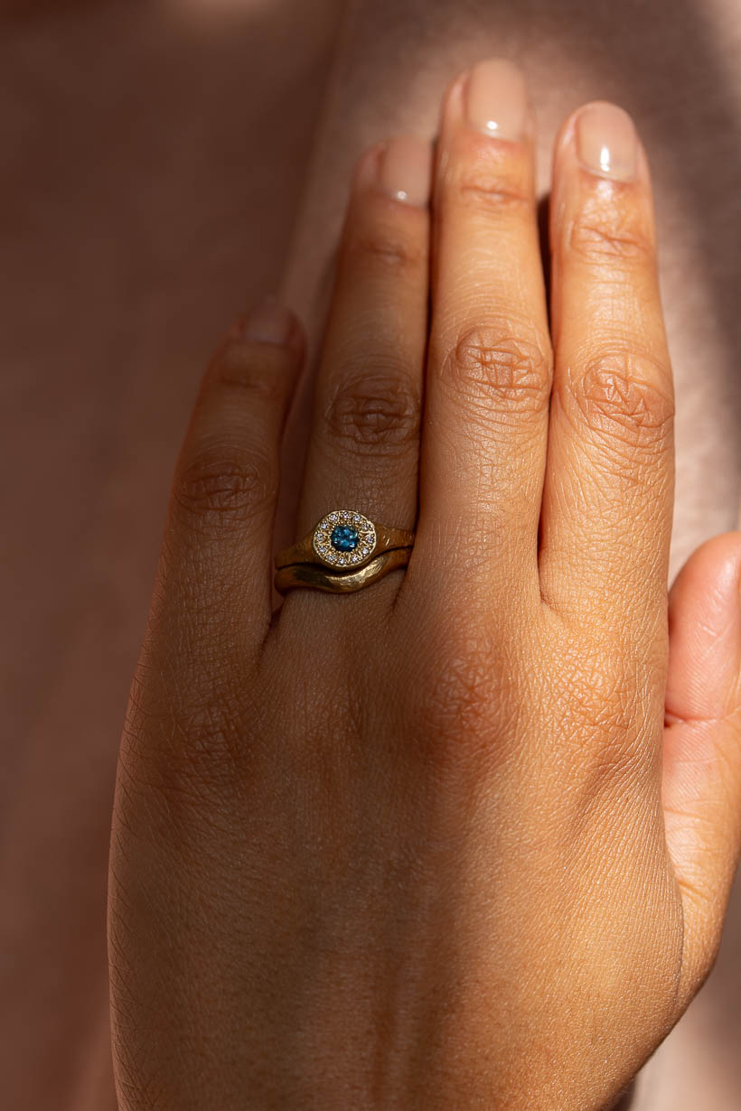 A close-up of a hand wearing a gold ring, the floral relic halo signet ring with a central round blue Australian sapphire surrounded by a halo of natural diamonds, worn with the 3mm wide Breeze ring, a curved, textured gold wedding band