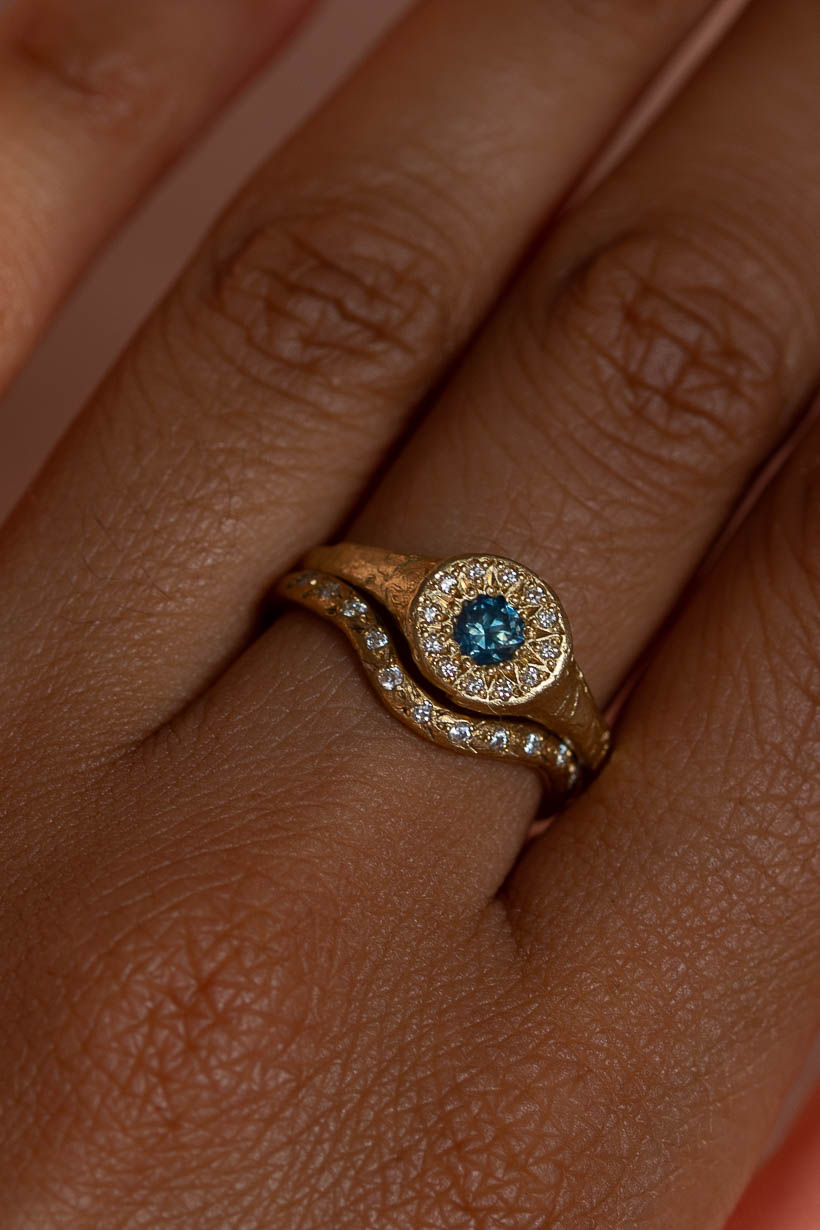 A close-up of a hand wearing a gold ring, the floral relic halo signet engagement ring, with a central round light blue Australian sapphire surrounded by a halo of natural diamonds with a wavy gold band, the rosette curved wedding band set with white natural diamonds. 