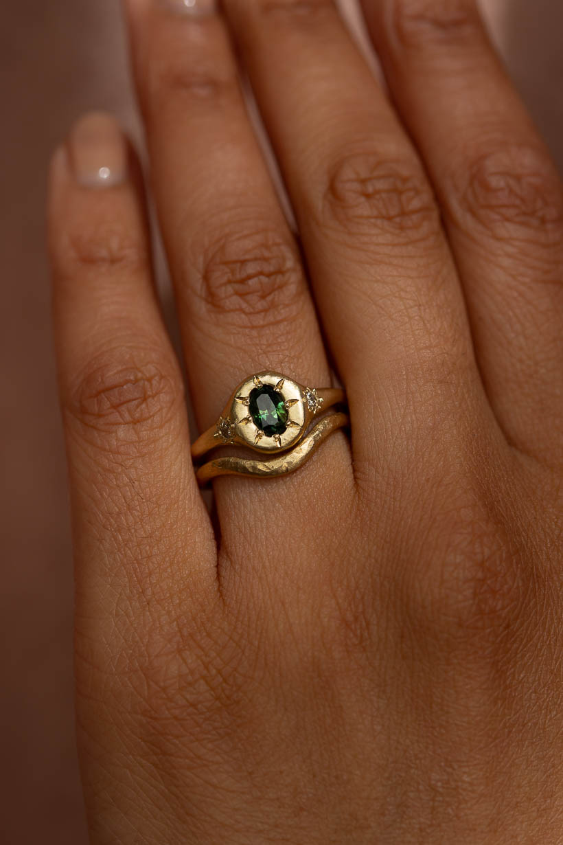 Close-up on hand of oval green sapphire with flower-set champagne diamonds in hand-textured gold. Stacked with curved 2mm Breeze wedding ring in yellow gold