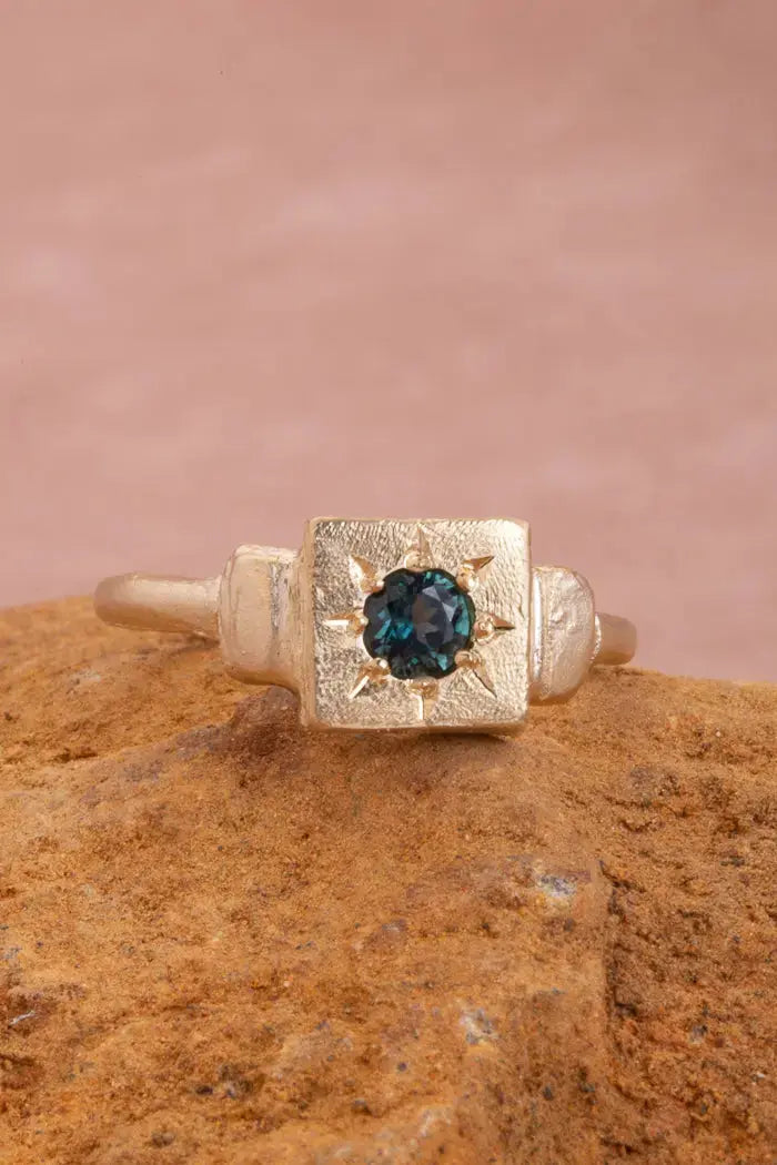 Front on photography of the dawn signet ring in gold. The teal topaz shines brightly and is star set in the recycled gold face. 